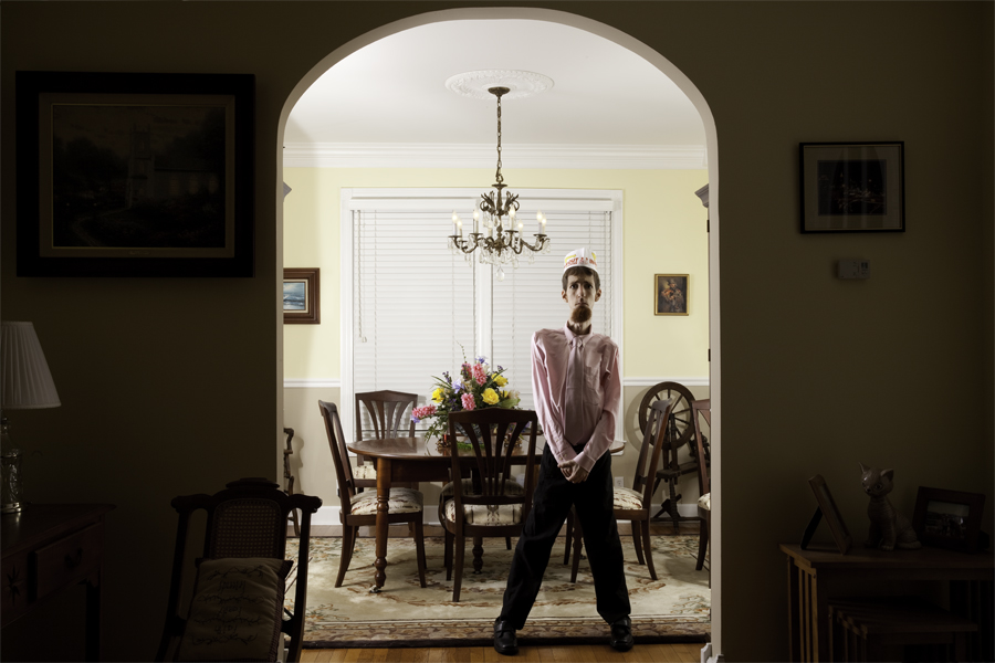 This Brian Charles Steel photo is a self-portrait of the artist in a dining room.  This image of Steel in the dining room was shot from the living room.  The dining room is well lit, but the living room is kept in shadow.  He is standing in front of the wooden dining room table with his hands folded neatly in front of him.  The table has a rug underneath it and a vase of flowers on top of it.  He is lit dramatically; both side so him are well lit, but his front is in shadow.  He is wearing black dress pants, pink dress shirt, and an In And Out cap.  His hair is short and brown.  He is framed by an archway between the living room and the dining room.  Above the table is a chandelier. Behind him on the left side is an antique spinning wheel.  On the wall behind him on the right there is an ocean painting.  In the living room on the right side there is a rocking chair and a lamp. On the left side is a table with picture frames and a ceramic cat.  
